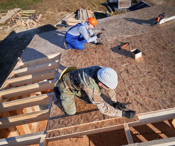 Carpenters hammering nail into OSB panel on the roof top of future cottage. Men workers building wooden frame house. Carpentry and construction concept.